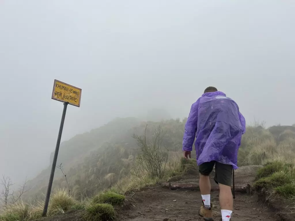  A person wearing a purple rain poncho is hiking up a misty trail. A yellow sign on the left indicates the destination “Khumai (45 mins)”. The trail is surrounded by grassy terrain and low visibility due to the fog.